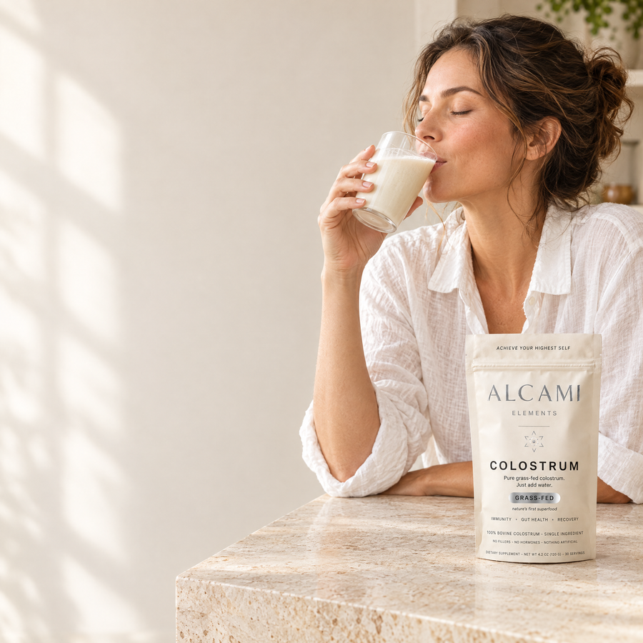 A woman sipping a creamy drink at a kitchen counter next to the Alcami Colostrum pouch