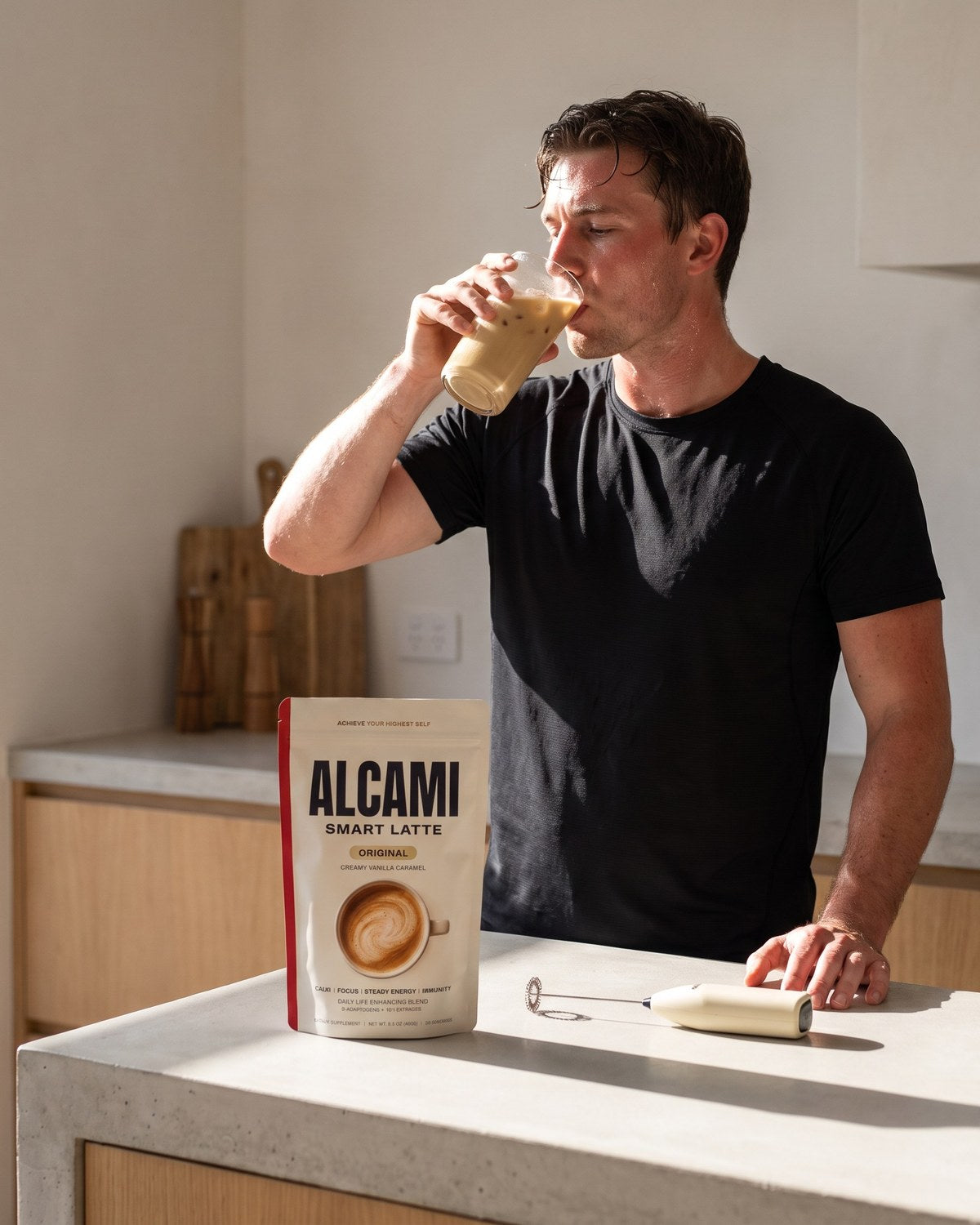 A man drinking an iced Alcami latte in his kitchen, the Smart Latte pouch and frother on the counter